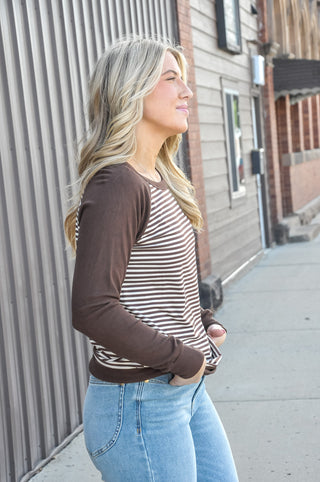This brown colorblock stripe raglan top pairs a brown and cream striped front panel with solid dark brown raglan sleeves and back for a laid-back baseball-inspired look that feels both effortless and elevated. 