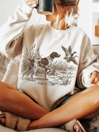 A woman sitting indoors wearing a cream sweatshirt with a black-and-white hunting dog and duck illustration on the front, holding a dark mug.