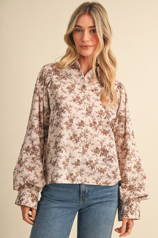A woman wearing a long-sleeve button-up blouse with a brown floral pattern on a light beige background, paired with blue jeans, against a solid beige studio backdrop.