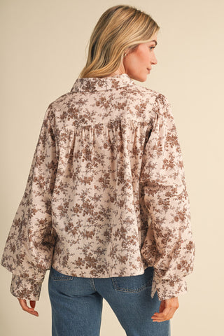 A woman shown from the back wearing a long-sleeve floral blouse with a gathered back and blue jeans, against a plain beige studio background.