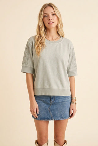 A woman modeling a sage green short-sleeve crewneck sweatshirt with textured knit sleeve panels, paired with a blue denim mini skirt and beige boots, against a solid cream background.