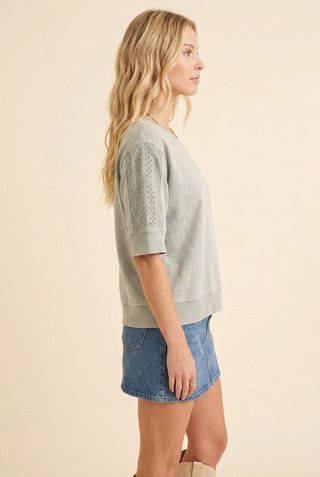 A woman shown in side profile wearing a light sage green short-sleeve knit top with textured eyelet lace sleeves and blue denim shorts, set against a solid beige studio background.