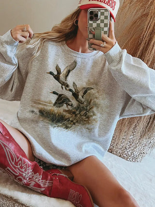A woman takes a mirror selfie wearing an oversized light gray heather crewneck sweatshirt with a graphic print of three ducks flying over marshland. She is also wearing red western boots and a red cap.